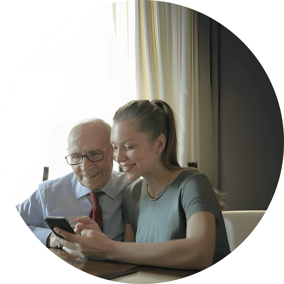 Homem idoso e mulher jovem sorrindo enquanto olham juntos para um celular, sentados à mesa em um ambiente iluminado por luz natural.